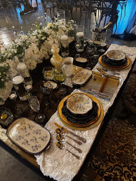 Luxury formal table setting with gold charger plates, patterned china, polished silverware, and white floral centerpiece on a black tablecloth and satin runner, styled for an upscale wedding or private dining event.