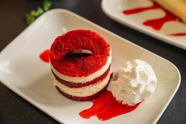 Red velvet layered dessert with cream filling, chocolate garnish, and whipped cream served on a white plate with red sauce drizzle.