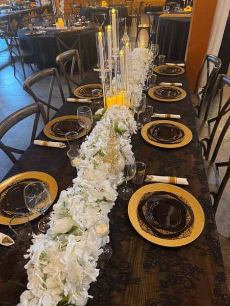 Elegant banquet dining table with black and gold place settings, amber glass plates, tall floating candles, and a lush white floral table runner, creating a sophisticated upscale event or wedding reception ambiance.