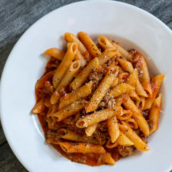 A plate of penne pasta is coated in a rich meat sauce, garnished with black pepper and grated cheese.