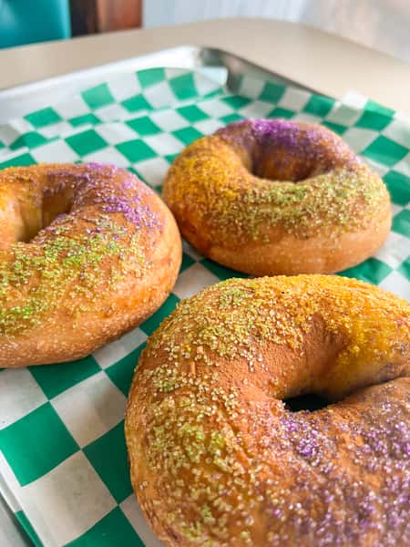 Three Mardi Gras bagels from Southbound Bagel and Coffee topped with purple, green, and gold sugar on checkered paper.