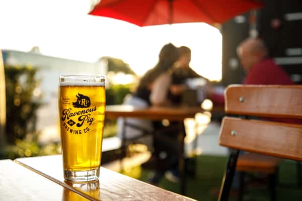 A pint of golden craft beer sits on a wooden outdoor table, glowing in the sunlight with a rich, foamy head. In the softly blurred background, guests relax on a patio beneath a bright red umbrella, creating a warm, inviting atmosphere perfect for enjoying drinks and casual dining outdoors.