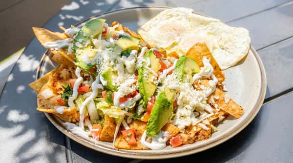 Chilaquiles with tortilla chips, avocado, lettuce, tomatoes, cheese, crema, and two fried eggs on a plate.