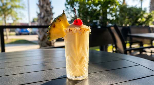 Yellow blended cocktail in a textured glass, garnished with a pineapple slice, cherry, and toasted coconut rim.