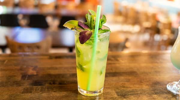 Mojito cocktail with mint, a lime wedge, purple basil, and a green straw in a glass on a wooden bar, blurry restaurant in background.