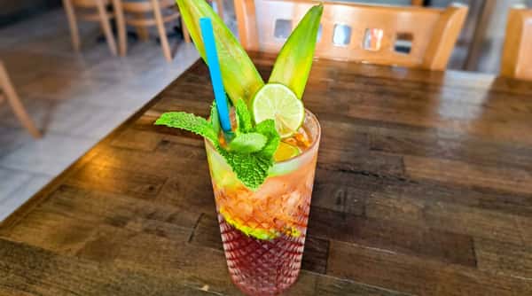 Tropical cocktail with pineapple leaves, mint, and lime garnish in a textured glass with a blue straw.