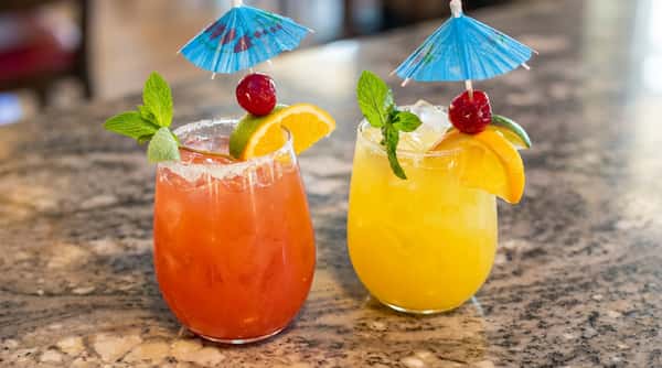Two colorful cocktails with sugared rims, garnished with citrus slices, cherries, mint, and cocktail umbrellas on a granite surface.