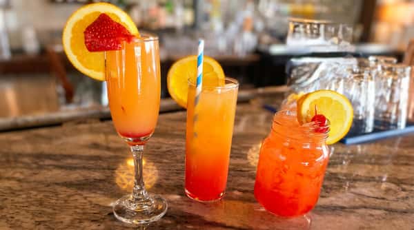 Three vibrant cocktails on a bar, garnished with orange slices, a strawberry, and a cherry.