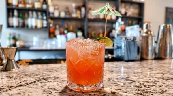 Orange cocktail in a textured glass with a salted rim, lime wedge, and green cocktail umbrella.