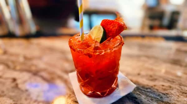 Red drink in a faceted glass with a striped straw, garnished with a lime wedge and strawberry, on a napkin.