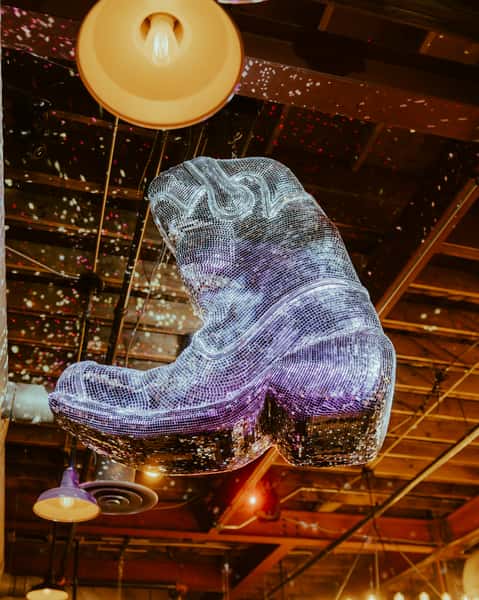 Oversized mirrored cowboy boot chandelier hanging above a vibrant Western-themed bar, creating a unique country nightlife centerpiece.