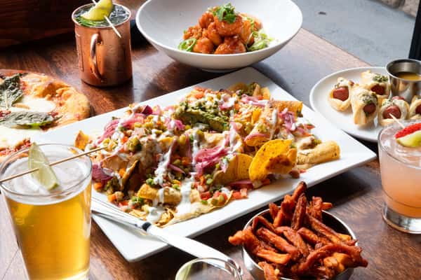 Shareable restaurant spread featuring loaded nachos, wood-fired pizza, boneless wings, sweet potato fries, cocktails, and craft beer for group dining.