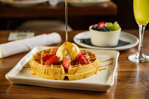 Belgian waffle topped with fresh strawberries, vanilla ice cream, and warm caramel drizzle, served with brunch cocktails and fresh fruit.