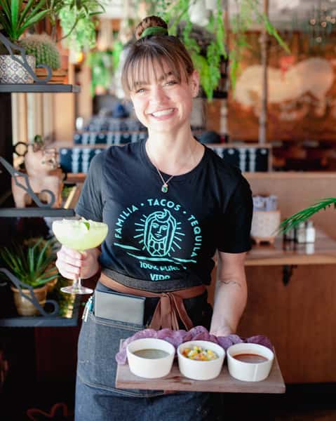 Restaurant server holding a handcrafted margarita and house-made sauces, showcasing warm hospitality in a vibrant Mexican-inspired dining room.