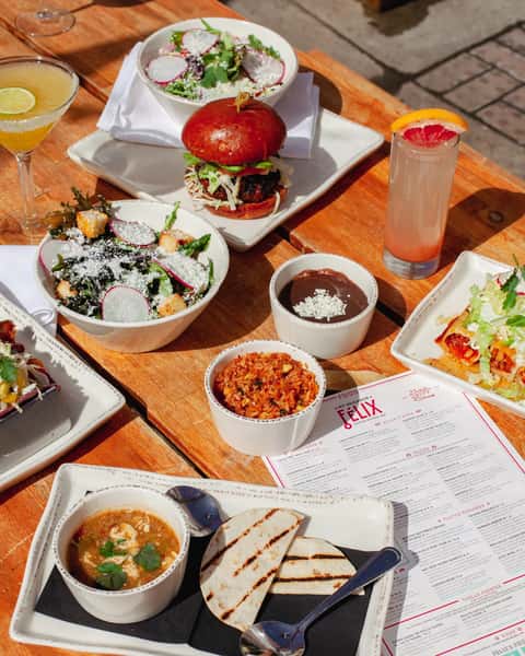 Outdoor dining table featuring gourmet burgers, fresh salads, house-made dips, cocktails, and Mexican-inspired dishes in a vibrant restaurant setting.