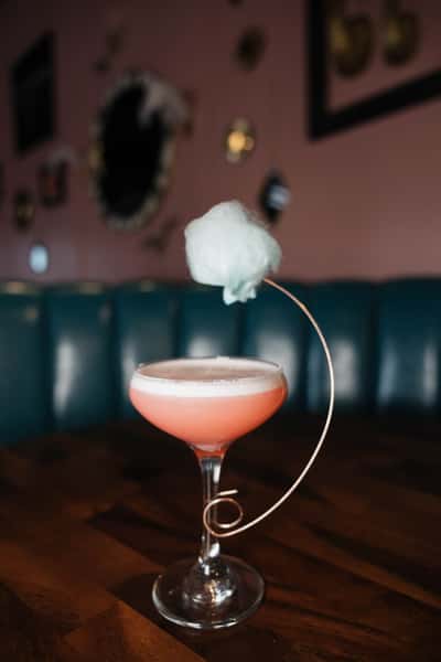 Signature pink cocktail topped with cotton candy, presented in an elegant coupe glass within a stylish contemporary cocktail lounge.