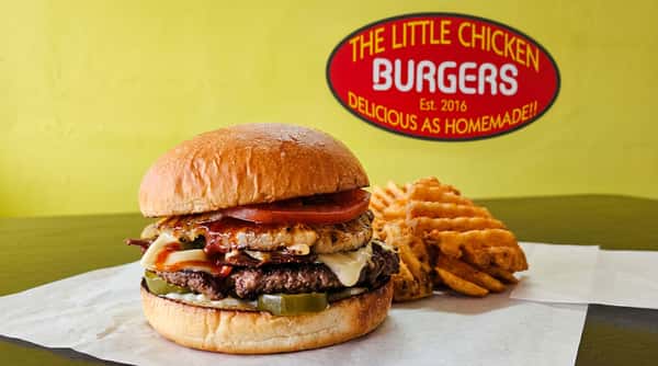 A double-patty hamburger with cheese, bacon, and pineapple, served with waffle fries on a white paper.