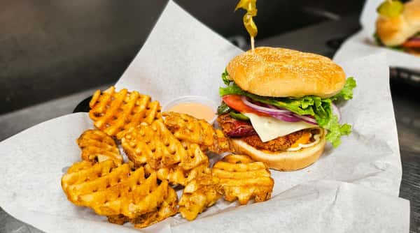 Fried chicken sandwich with bacon, pepper jack cheese, red onion, lettuce, tomato, and a pepper skewer, served with waffle fries and a side of sauce on white paper.