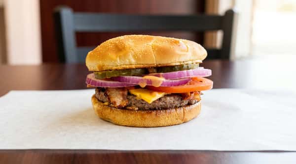 Bacon cheeseburger with red onion, tomato, pickles, and sauce on a sesame seed bun, on a white paper liner.