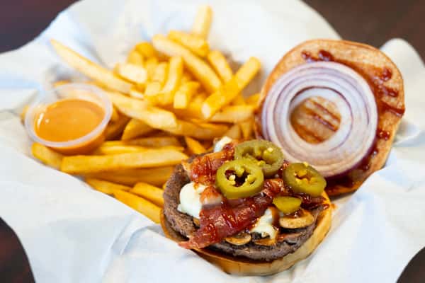 An open hamburger with beef patty, bacon, jalapenos, mushrooms, and BBQ sauce served with a pile of golden fries and dipping sauce in a white basket.