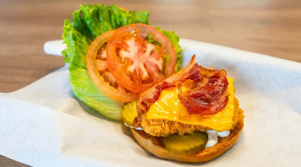 Open-face fried chicken sandwich with melted cheese, bacon, pickles, lettuce, and tomato on a white paper-lined tray.