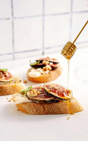 Toasted bread topped with fresh figs, cheese, walnuts, and mint as honey drizzles from a dipper onto the front slice.
