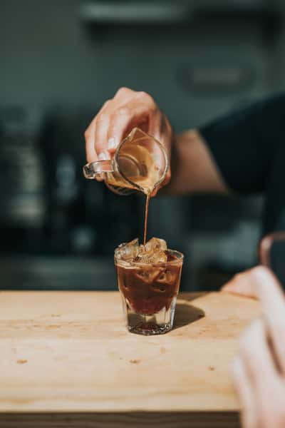 Espresso being poured from a small glass pitcher over ice in a clear glass, creating an iced coffee drink on a wooden counter.