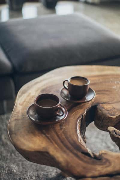 Two cups of coffee sit on a rustic, natural wood table with a raw edge design, in front of a cozy, dark-colored sofa.