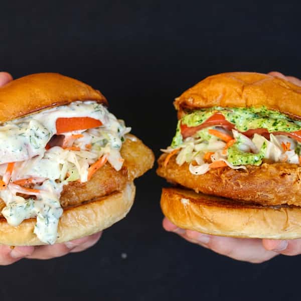 Two crispy fried chicken sandwiches topped with creamy herb sauce, fresh coleslaw, and tomatoes on toasted brioche buns, held side by side against a dark background.