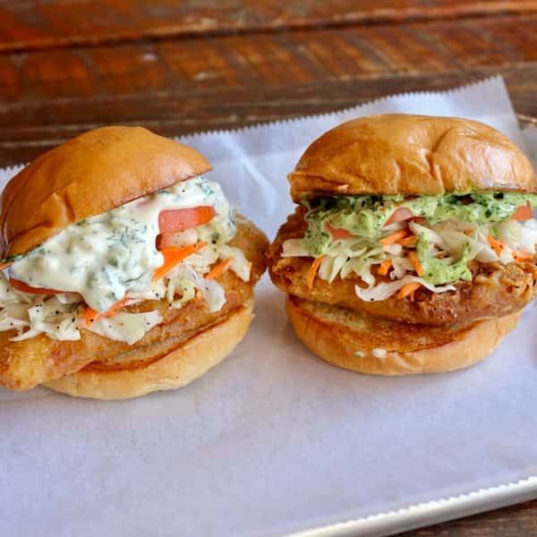 Two crispy fried chicken sandwiches topped with creamy herb sauce, fresh coleslaw, and tomatoes on toasted buns, served on parchment paper at Social Burger.