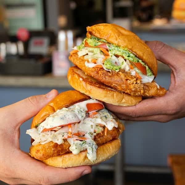 Two crispy fried chicken sandwiches topped with creamy herb sauce, fresh coleslaw, tomatoes, and toasted brioche buns, held by customers inside Social Burger.