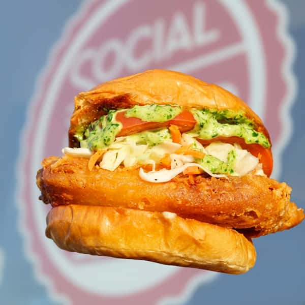 Crispy fried chicken sandwich topped with creamy herb sauce, fresh coleslaw, and sliced tomatoes in front of the Social Burger restaurant storefront.