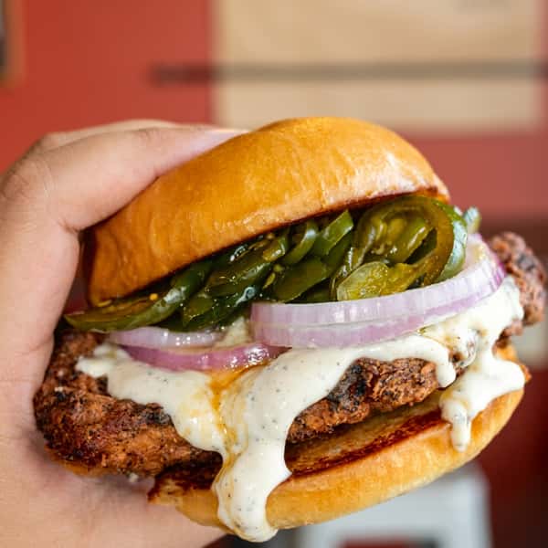 Hand holding a grilled chicken sandwich with melted cheese, jalapeños, red onions, and creamy garlic sauce on a toasted brioche bun at Social Burger.