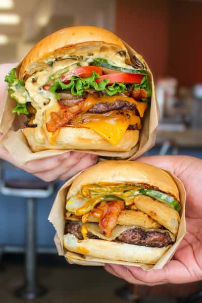 Two handcrafted Social Burger gourmet burgers loaded with bacon, cheddar cheese, grilled onions, fresh vegetables, and crispy onion rings, held by customers inside the restaurant.