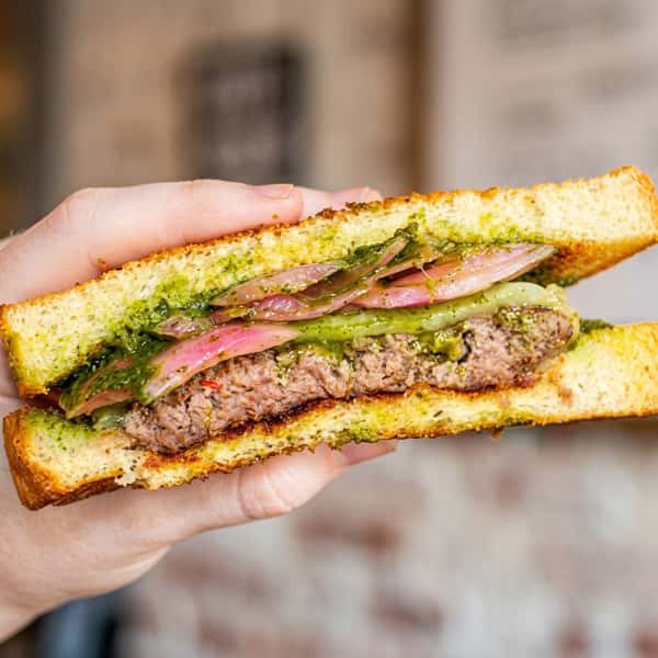 Hand holding a Social Burger garlic toast burger with a juicy beef patty, avocado, pickled red onions, and herb sauce in a close-up cross-section.