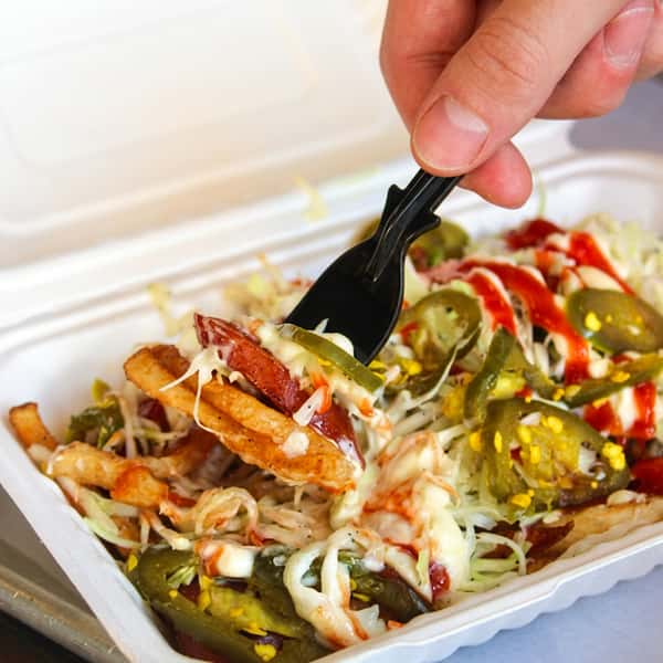 Loaded seasoned fries topped with melted cheese, bacon, jalapeños, shredded lettuce, and signature sauce in a takeout container.