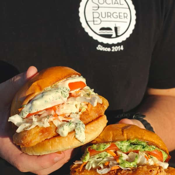 Two crispy fried chicken sandwiches topped with creamy herb sauce, fresh coleslaw, and tomatoes, held by a Social Burger team member wearing a branded restaurant shirt.