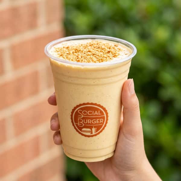 Hand holding a creamy Social Burger milkshake topped with cookie crumbs, served in a branded cup with a fresh outdoor background.