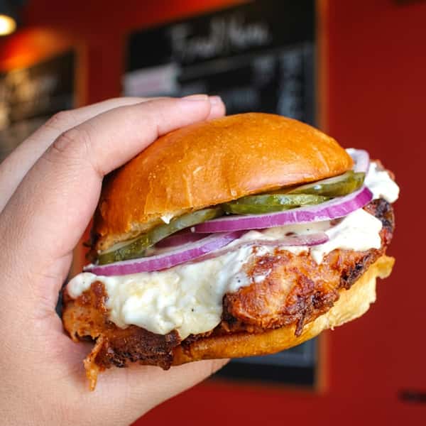 Hand holding a grilled chicken sandwich with melted cheese, pickles, red onions, and creamy sauce on a toasted brioche bun inside Social Burger.
