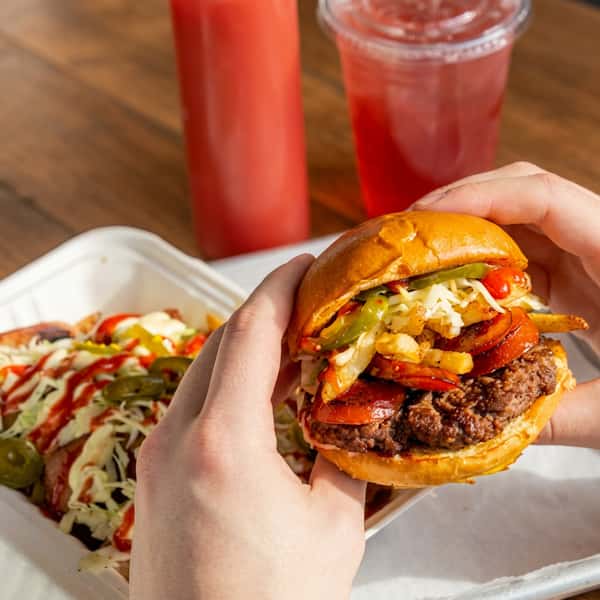 Hands holding a loaded Social Burger topped with bacon, fries, jalapeños, and shredded lettuce, served with loaded fries and a refreshing drink.