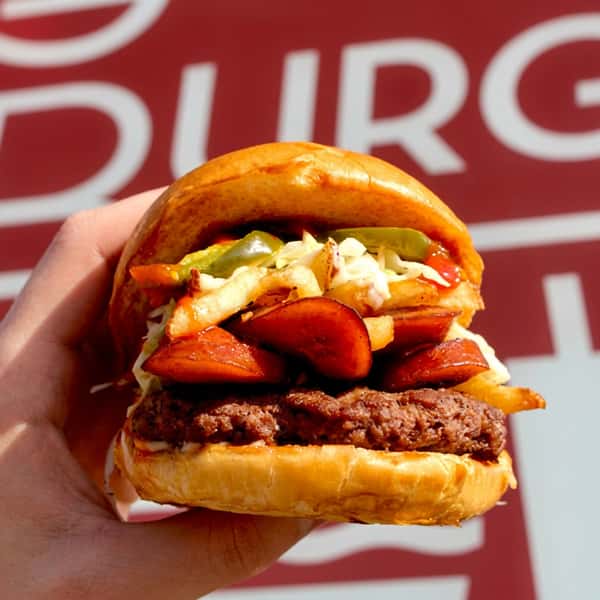 Hand holding a loaded Social Burger with grilled beef, sliced sausage, fries, jalapeños, coleslaw, and fresh toppings in front of the restaurant.