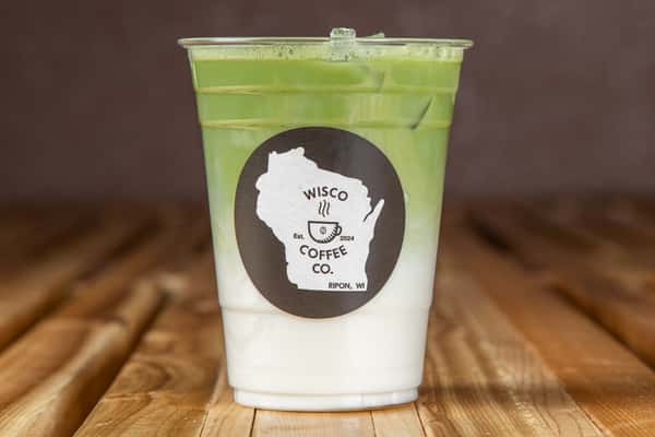Iced matcha latte with layered green tea and milk in a clear plastic cup, featuring a minimalist logo, set on a rustic wooden table in a modern café setting.