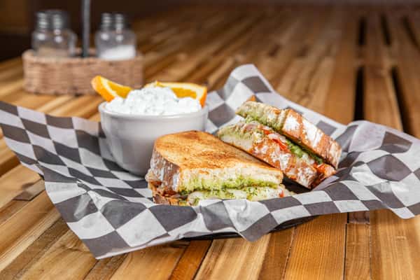 Toasted tomato pesto grilled cheese sandwich with melted cheese and fresh tomato slices on golden-brown bread, served with orange slices and a creamy side on checkered paper, highlighting a cozy café meal.