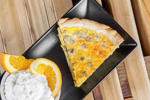 Savory slice of quiche with a golden, flaky crust and cheesy egg filling, served on a black plate with a side of whipped cream and orange garnish, set on a rustic wooden table in a cozy café setting.