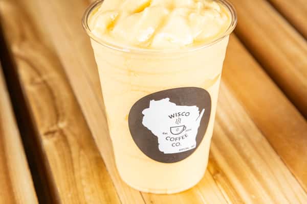 Creamy mango or tropical fruit smoothie in a clear plastic cup with a minimalist café logo, served over ice on a wooden table, showcasing a bright and refreshing drink.