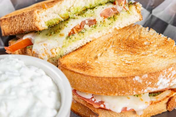 Toasted tomato pesto grilled cheese sandwich with melted cheese, fresh tomato slices, and basil pesto on golden-brown bread, served in a basket on checkered paper, capturing a warm and savory café-style comfort food.