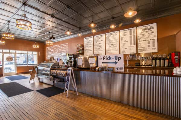 Coffee shop counter area with pastry display, pick-up station, and barista workspace, featuring warm lighting and industrial-style design for an inviting café experience.