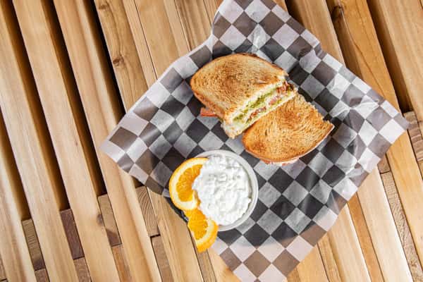 Toasted tomato pesto grilled cheese sandwich with melted cheese and fresh tomato slices on golden-brown bread, served with orange slices and a creamy side on checkered paper, highlighting a cozy café meal.