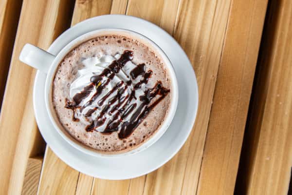 Rich hot chocolate with chocolate syrup drizzle and frothy foam in a white cup, served on a wooden table, highlighting a warm café dessert drink.