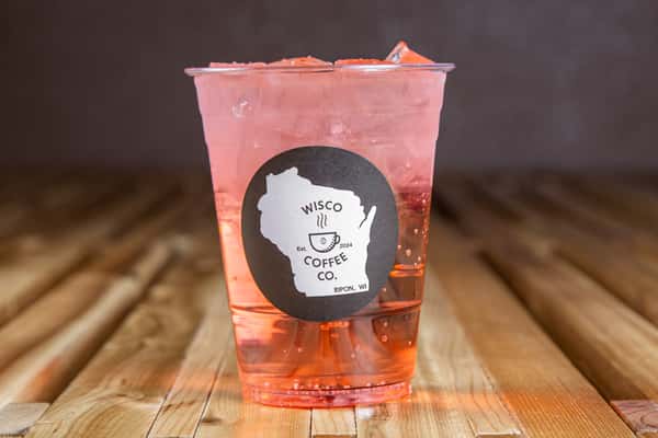 Refreshing watermelon lemonade or fruit refresher with a light pink hue, served over ice in a clear plastic cup with a minimalist café logo, set on a rustic wooden table for a vibrant summer drink presentation.